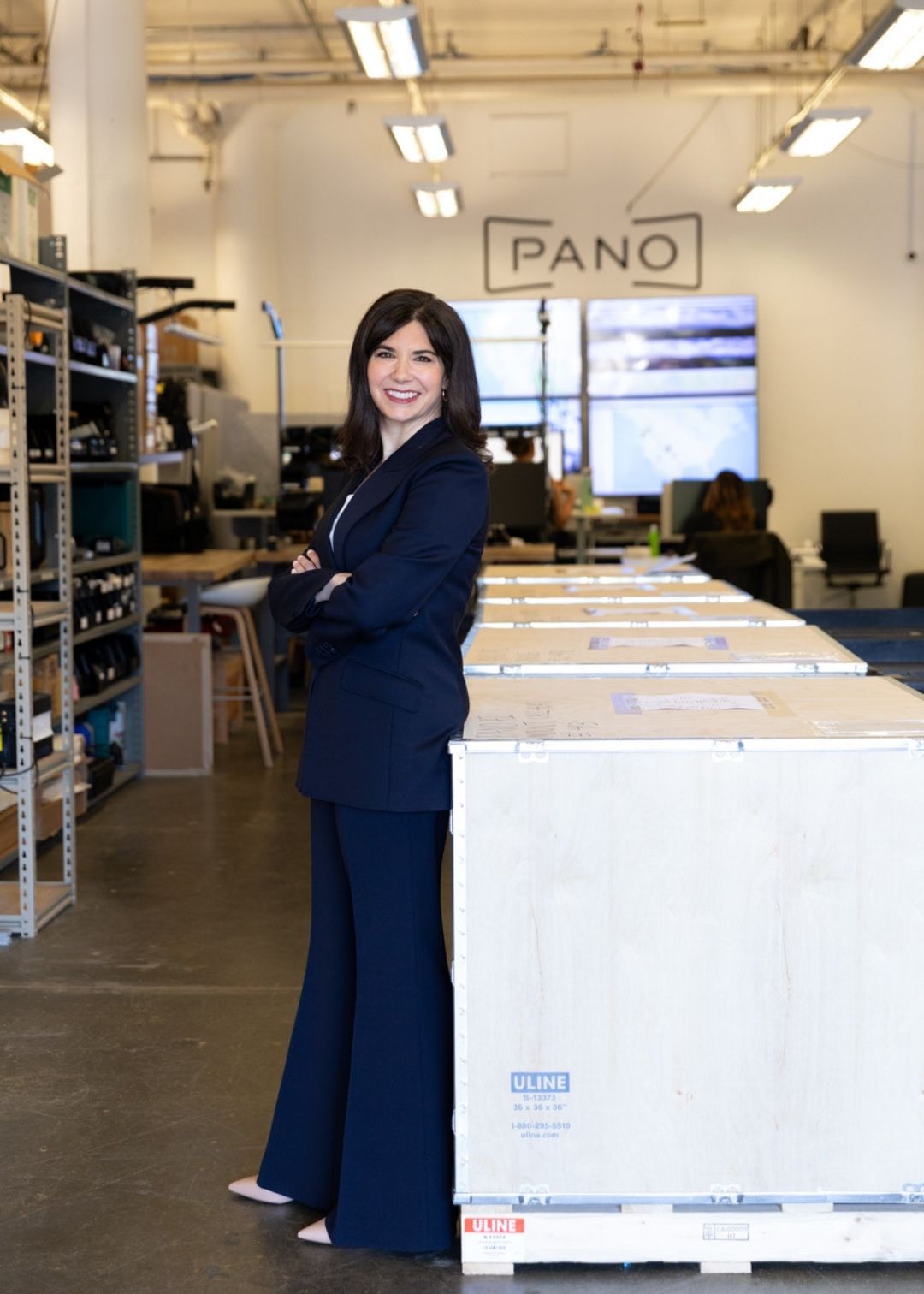 A woman in a dark suit stands with arms crossed next to large shipping crates in an industrial office space with a "PANO" sign and testimonials displayed in the background.